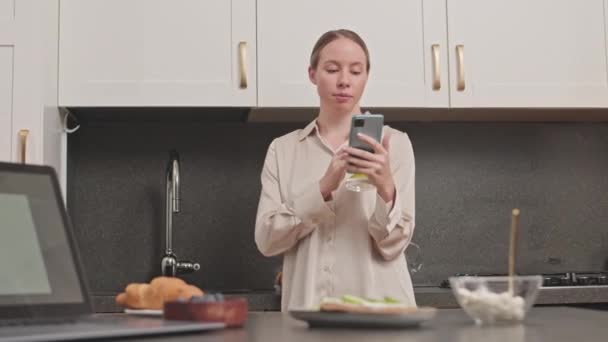 Medium laukaus nuori kaunis valkoihoinen nainen juo lasia sitruunavettä ja käyttää älypuhelin seisoo moderni keittiö yllään silkki pyjama paita - Materiaali, video
