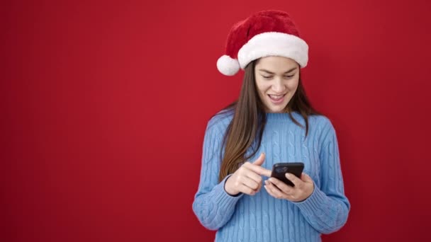 Νεαρή καυκάσια γυναίκα που φοράει χριστουγεννιάτικο καπέλο χρησιμοποιώντας smartphone κοιτάζοντας στο πλάι πάνω από απομονωμένο κόκκινο φόντο - Πλάνα, βίντεο