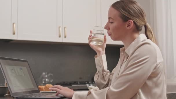 Side view taille omhoog slowmo van jonge blanke vrouw werken op laptop terwijl zitten aan keukentafel in modern appartement overdag - Video