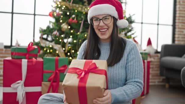 Joven mujer hispana sonriendo usando sombrero de navidad sosteniendo presente en casa - Imágenes, Vídeo