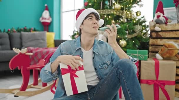 Hombre hispano joven sosteniendo regalo de Navidad enviando mensaje de voz por teléfono inteligente en casa - Metraje, vídeo