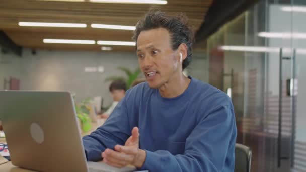 Medium shot van Aziatische man in draadloze oortelefoons spreken via videogesprek op laptop tijdens de dag in de co-working room - Video