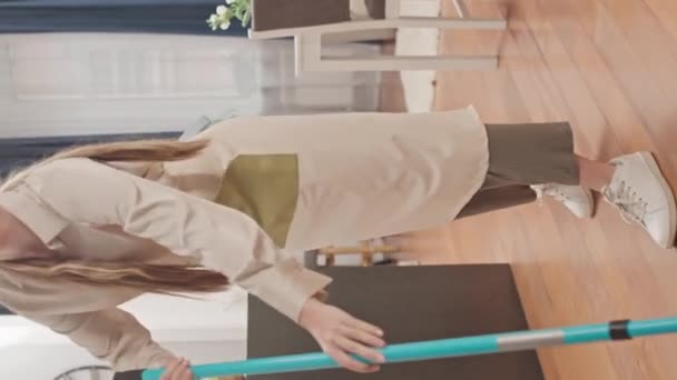 Вертикальный снимок молодой кавказки в фартуке, вытирающей пол в светлой современной однокомнатной квартире - Кадры, видео
