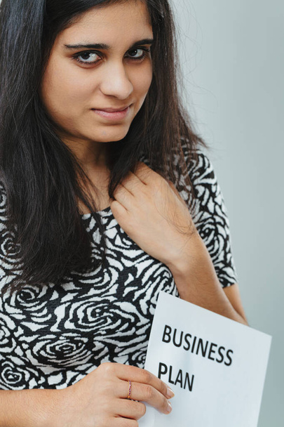 С решительным взглядом темноволосая женщина хватается за "бизнес-план". Замысловатый розовый узор на ее одежде выделяется, заставляя ее выглядеть как профессионально, так и стильно - Фото, изображение