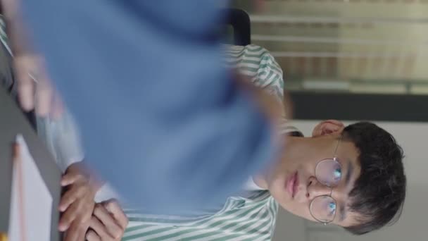 Vertical shot of gen Z Asian man in striped shirt and breglasses sitting at meeting table in office and having a conversation with fellow ague - Záběry, video
