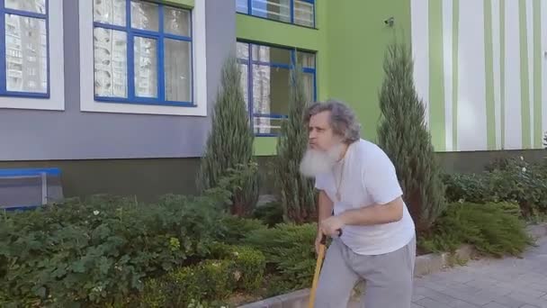 El viejo abuelo monta un monopatín por la calle. El viejo ayuda con un bastón para montar un monopatín. Un anciano está aprendiendo a montar un monopatín. - Metraje, vídeo