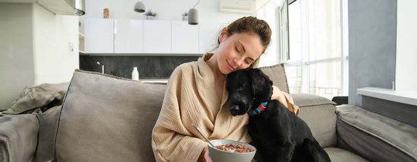 Kobieta z krótkimi włosami siedząca na kanapie w domu, jedząca płatki na śniadanie i głaskająca swojego ukochanego psa. Pies wącha miskę płatków.. - Zdjęcie, obraz