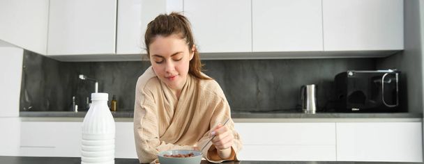 Πορτρέτο της όμορφης γυναίκας στο σπίτι, τρώγοντας το πρωινό της, κρατώντας κουτάλι, ακουμπώντας στον πάγκο εργασίας και έχοντας δημητριακά με γάλα. - Φωτογραφία, εικόνα