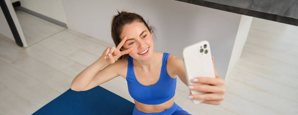 Portret pięknej atletki, kobiety ćwiczącej w domu na gumowej macie do jogi, robi selfie na smartfonie, robi zdjęcia do mediów społecznościowych instruktora fitness. - Zdjęcie, obraz