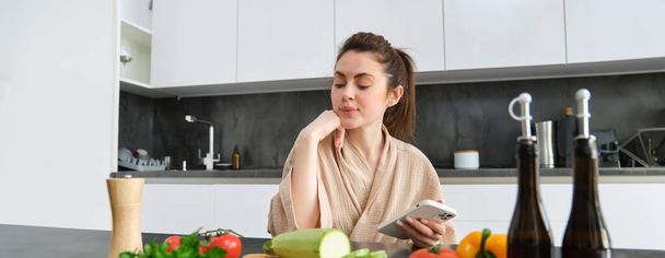 Portrait of young woman ordering groceries on smartphone app, holding tomato, sitting near chopping board with vegetables. Girl looking for recipe online, using mobile phone. - Photo, Image