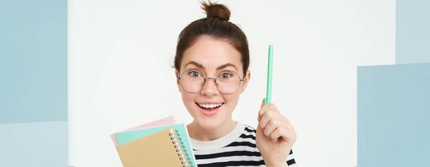 Πορτρέτο της γυναίκας σε γυαλιά, δάσκαλος σηκώνει το χέρι με στυλό, eureka χειρονομία, έχει ιδέα ή λύση, κρατά σημειωματάρια έγγραφα, στέκεται πάνω από το λευκό φόντο. - Φωτογραφία, εικόνα