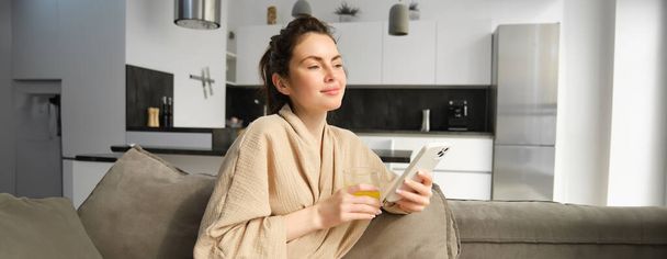 Красивая молодая женщина отдыхает на диване, наслаждаясь утром дома, держа стакан апельсинового сока и смартфон, глядя в окно. - Фото, изображение