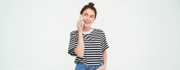 Joyeux jeune femme parle sur un téléphone mobile, discute sur le téléphone, utilise un smartphone, se tient sur fond blanc. Espace de copie - Photo, image