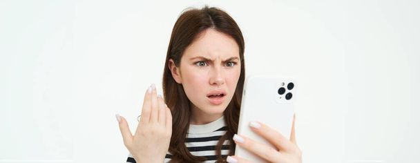 Portret zdezorientowanej, wściekłej kobiety marszczącej brwi, czytającej tekst na smartfonie, oglądającej wideo na telefonie komórkowym z zaszokowaną, zakłopotaną twarzą, odizolowanej na białym tle. - Zdjęcie, obraz