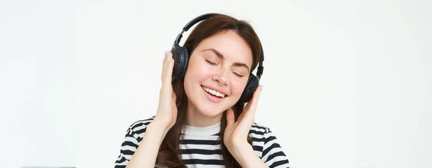 Porträt einer schönen Frau mit drahtlosen Kopfhörern, Musik hörend, Kopfhörer benutzend, in die Kamera lächelnd, vor weißem Hintergrund stehend. - Foto, Bild