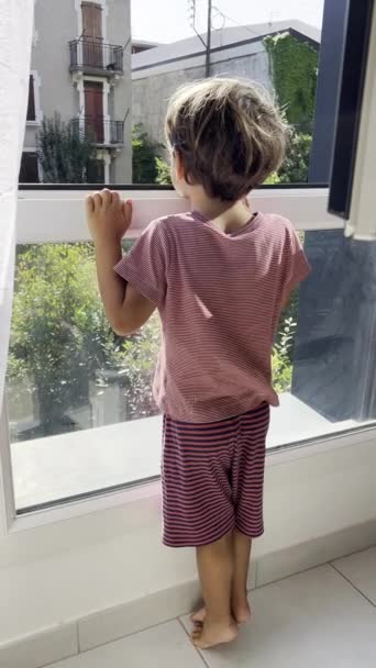 Niño curioso en pijama mirando a través de la ventana para espiar el vecindario - Imágenes, Vídeo