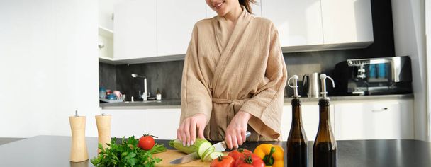 Zdrowy styl życia. Młoda kobieta w szlafroku przygotowuje jedzenie, kroi warzywa, gotuje obiad na blacie kuchennym, stoi na białym tle. - Zdjęcie, obraz