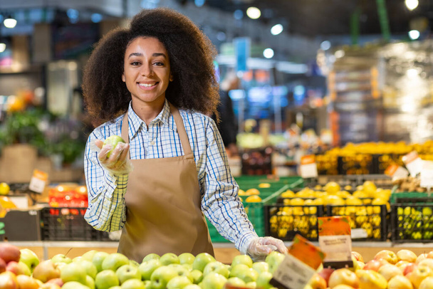 Retrato de una joven vendedora en un supermercado, tienda de comestibles, una vendedora sonriente mirando a la cámara con satisfacción, mirando manzanas, una mujer hispana con el pelo rizado en un delantal. - Foto, Imagen