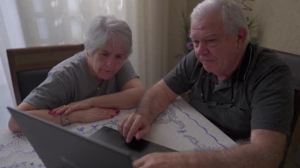Coppia anziana che interagisce con il computer portatile in salotto, marito che indica contenuti online alla moglie - Filmati, video