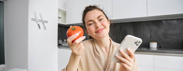 Primer plano retrato de una hermosa mujer sonriente, sosteniendo tomate fresco, sentado en la cocina con teléfono inteligente, ordena verduras en línea, utilizando la aplicación para comprar comestibles, utilizando el teléfono móvil. - Foto, Imagen
