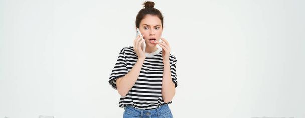 Image of woman with shocked face, looks confused while chats over the phone, listens to person on telephone, white background. - Photo, Image
