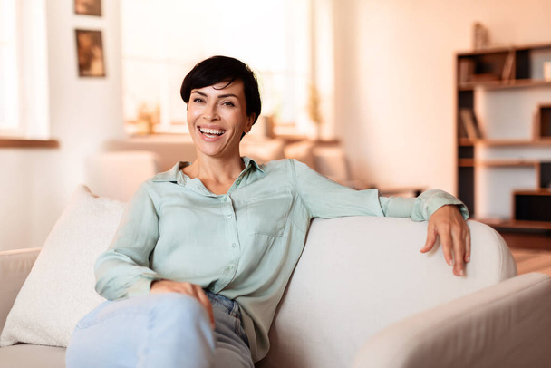 Женщина средних лет с темными волосами улыбается, глядя на камеру, позирующую на диване в гостиной дома. Привлекательная женщина в повседневной жизни отдыхает в уютном внутреннем интерьере на выходных - Фото, изображение