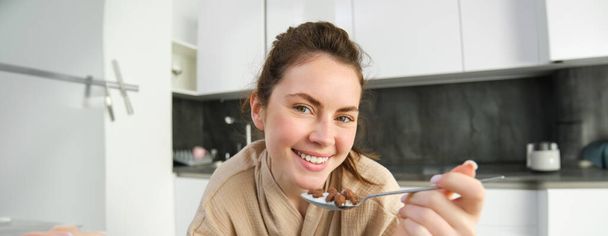 Retrato de una hermosa mujer en casa, desayunando, sosteniendo cuchara, apoyándose en la encimera y comiendo cereales con leche. - Foto, imagen