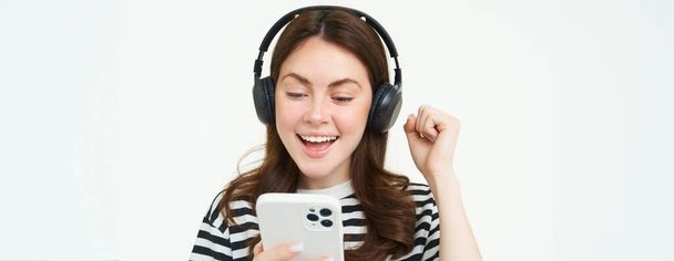Χαρούμενη, όμορφη νεαρή γυναίκα που χρησιμοποιεί την εφαρμογή smartphone, γιορτάζει, δείχνει χαρούμενη ενώ παίζει με το κινητό τηλέφωνο, ακούει μουσική σε ασύρματα ακουστικά. - Φωτογραφία, εικόνα