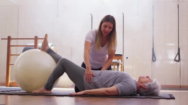 Physiotherapist Guiding Elderly Woman in Pilates Ball Exercise, Senior Lady Engaged in Floor Workout Routine with Coach Assistance - Felvétel, videó