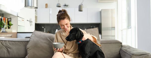 Σπίτι και τρόπος ζωής έννοια. Όμορφη νεαρή γυναίκα με σκύλο, κάθεται στον καναπέ, τρώει δημητριακά και αγκαλιάζει το κουτάβι της, το κορίτσι τρώει πρωινό με μπουρνούζι. - Φωτογραφία, εικόνα