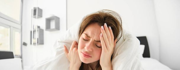 Portret van een vrouw met hoofdpijn, liggend in bed en het aanraken van het hoofd met gefrustreerd, fronsend gezicht, voelt zich niet goed, blijft in de slaapkamer als met migraine in de ochtend. - Foto, afbeelding