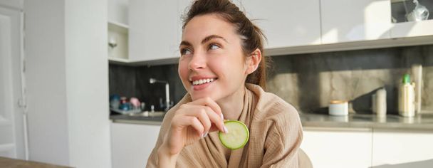 Retrato de una joven feliz y sonriente en la cocina, cocinando, picando calabacín, sosteniendo verduras y luciendo feliz, preparando comida vegana en casa. - Foto, Imagen