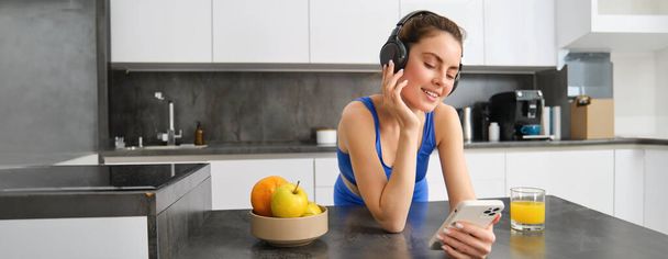 Зображення щасливої, стильної молодої спортивної жінки, що стоїть на кухні та п'є апельсиновий сік, слухає музику в навушниках, використовуючи додаток для смартфона. - Фото, зображення