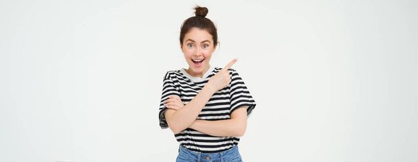 Image of excited young woman, smiling, showing amazing price discounts, pointing left, demonstrating something awesome, standing over white background. - Photo, Image