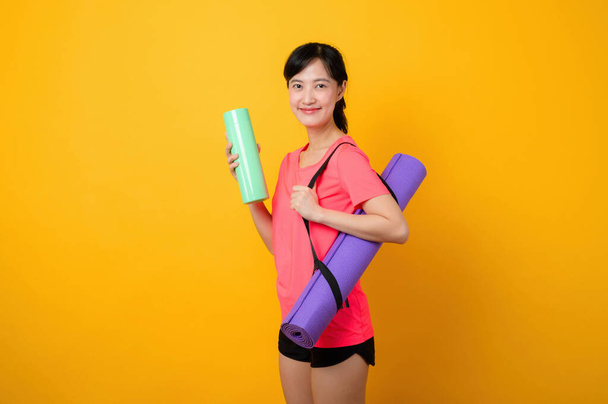 Портрет азіатської молодої спортивної фітнес-жінки щаслива посмішка в рожевому спортивному одязі та йога-матусі робить тренувальні тренування на жовтому студійному фоні. здорового способу життя. - Фото, зображення