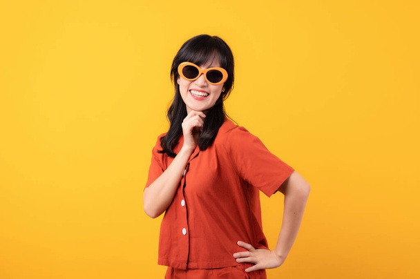 Πορτρέτο όμορφη νεαρή Ασιάτισσα γυναίκα χαρούμενο χαμόγελο ντυμένο με πορτοκαλί ρούχα και γυαλιά ηλίου αναζητούν γιορτάζουν με το χέρι στο πρόσωπό της απομονώνονται σε κίτρινο φόντο στούντιο. - Φωτογραφία, εικόνα