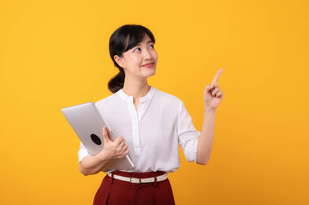 Portret młoda piękna azjatycka kobieta przedsiębiorca szczęśliwy uśmiech noszenie białej koszuli i czerwone rośliny wskazując gest palca do wolnej przestrzeni kopii i gospodarstwa laptop izolacji na żółtym tle. - Zdjęcie, obraz