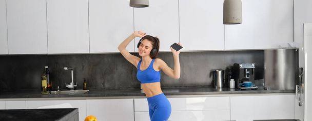 Πορτρέτο της όμορφης blogger φυσικής κατάστασης, γυναίκα με ακουστικά, ακούγοντας μουσική και χορεύοντας στην κουζίνα, φορώντας μπλε κολάν και αθλητικά παπούτσια. - Φωτογραφία, εικόνα