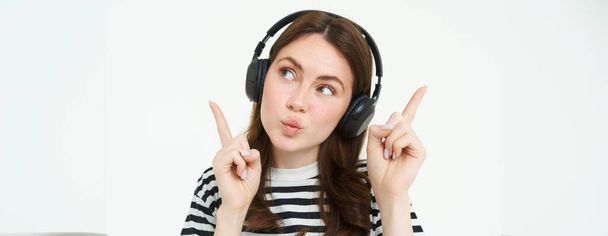 Πορτρέτο της νεαρής μελαχρινής γυναίκας στα ακουστικά, ακουστική μουσική στα ακουστικά, δείχνοντας τα δάχτυλα προς τα πάνω, δείχνει πανό, διαφήμιση, απομονώνονται σε λευκό φόντο. - Φωτογραφία, εικόνα