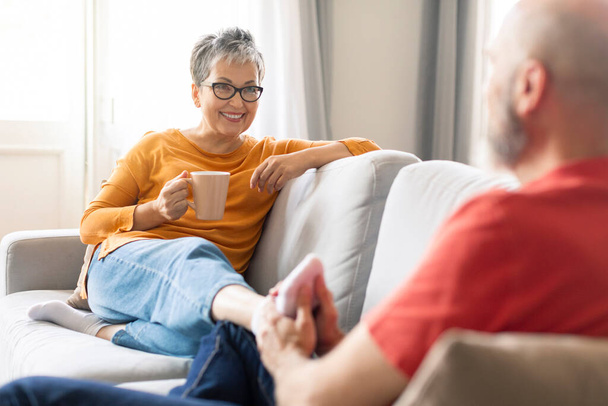 Любящий старший муж делает массаж ног своей жене дома, помогает ей расслабиться, счастливые пожилые супруги отдыхают на диване в гостиной вместе, женщина пьет кофе, избирательный фокус - Фото, изображение