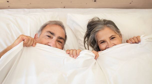 Smiling old caucasian man and lady wakes up in bed, enjoy good morning together, have fun under blanket in bedroom, top view. Weekend and spare time, relationship and love, lifestyle, relax - Photo, Image