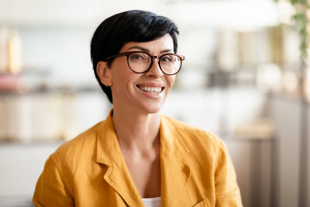 幸せな中年の女性の肖像画は,屋内カメラで楽観的に微笑み,黄色いジャケットを着ています. 現代オフィスの眼鏡でポーズショートヘアカットのブルネット女性起業家 - 写真・画像