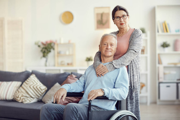 Portrait of content attractive wife in eyeglasses embracing husband in wheelchair around neck while helping him in recovery - Φωτογραφία, εικόνα