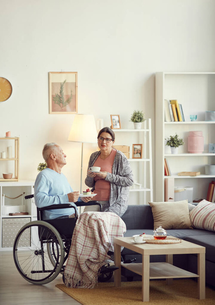 Serious mature woman in eyeglasses sitting on sofa in living room and holding cup of tea while talking to husband in wheelchair - Photo, image