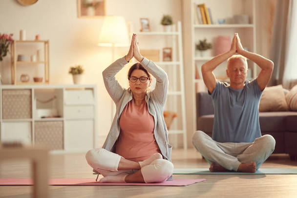 Serious senior couple focused on thoughts sitting with crossed legs and raising hands in Namaste gesture up while meditating at home - Foto, Bild