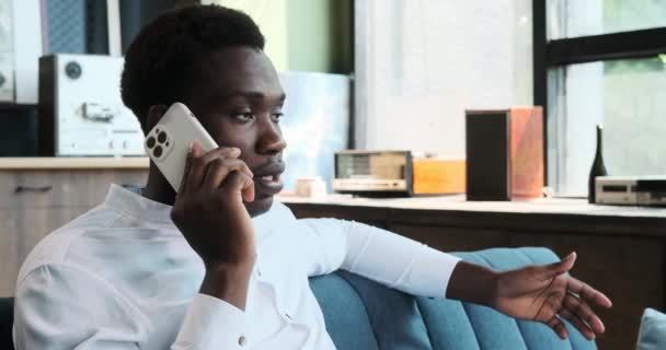 Un uomo di colore si impegna in una conversazione telefonica mentre è seduto sul divano in soggiorno. La sua comunicazione focalizzata è catturata in questa scena della vita quotidiana. - Filmati, video