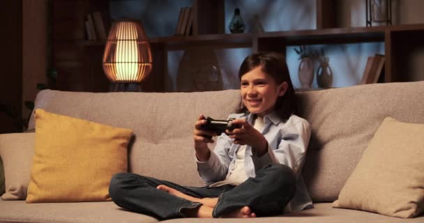Iskolásfiú ül a kanapén, teljesen lefoglal egy videojáték, kezek ügyesen manőverező a gamepad. A szemei a képernyőre vannak rögzítve, és az arca koncentrációt és izgalmat mutat.. - Felvétel, videó