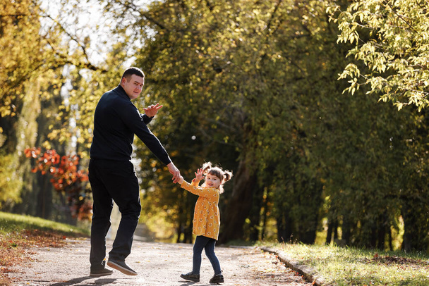 Happy fathers day. Dad holds hand his little daughter, walks, waves hands at camera outdoors at autumn park. Single daddy with child relaxes and spends time together. Family day and childhood concept - Photo, Image