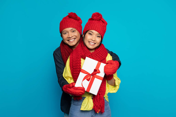 Happy millennial asian guy in hat show box present and hugs woman, isolated on blue studio background. Weekend, cold, holidays, winter and New Year celebration together, Xmas gifts - Photo, Image
