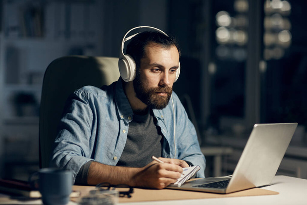 Serious concentrated hipster young bearded motion designer in headphones sitting at table and drawing in sketchpad while working till late evening - Фото, зображення
